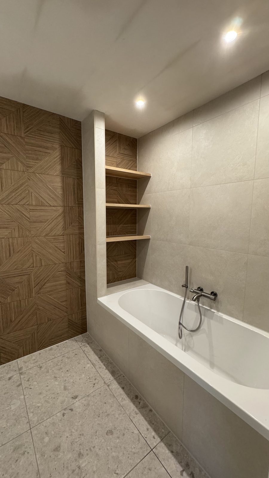 Badkamer met ligbad, wand met houtlooktegels, nisplanken en terrazzo vloer