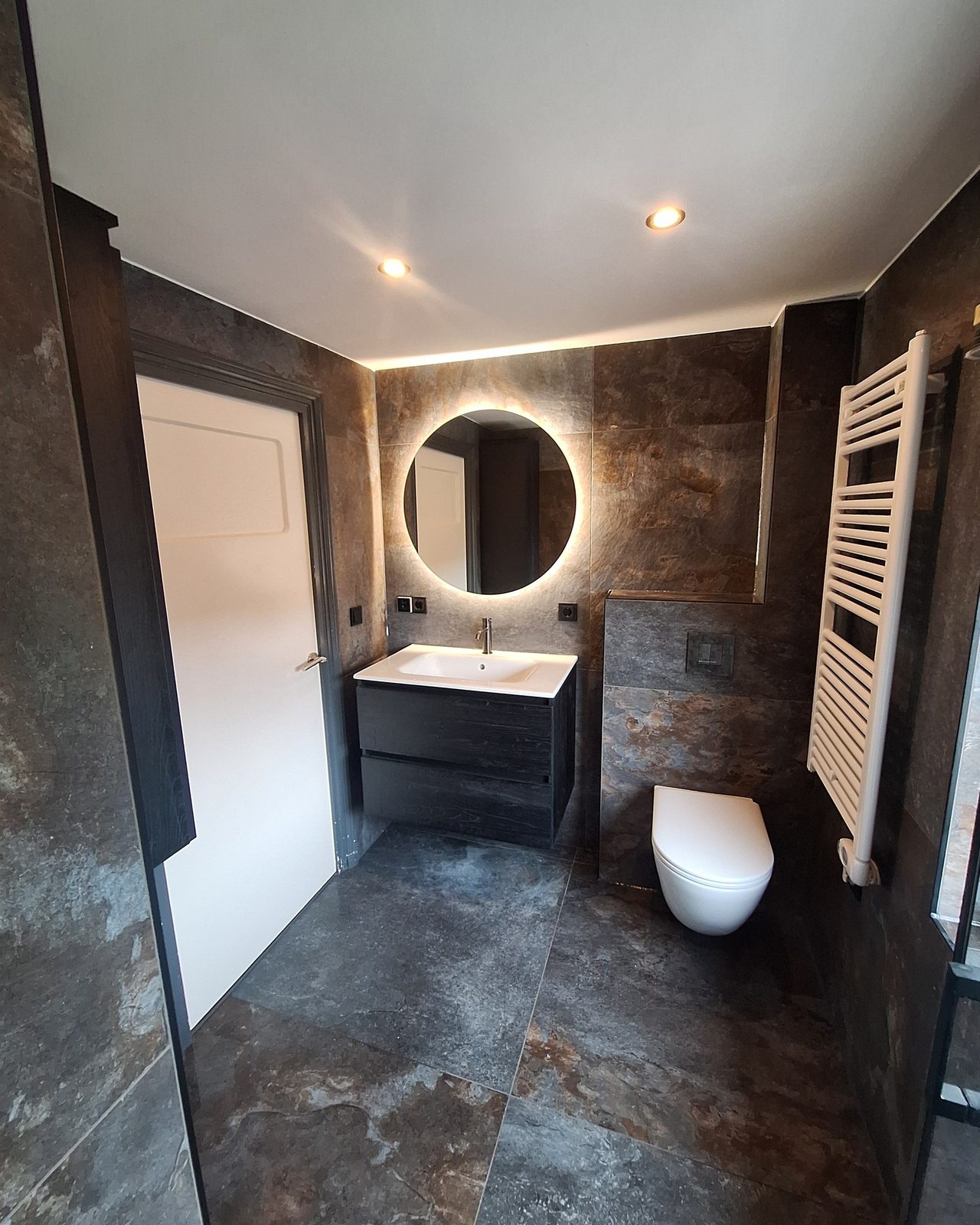 Badkamer met donkere tegels in roestlook, ronde spiegel en zwevend toilet